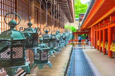 A row of ornate metal lanterns hang beside a wooden building with sliding doors, opposite a corridor with bright red columns at a traditional Japanese shrine. A few people walk in the background under lush green trees.