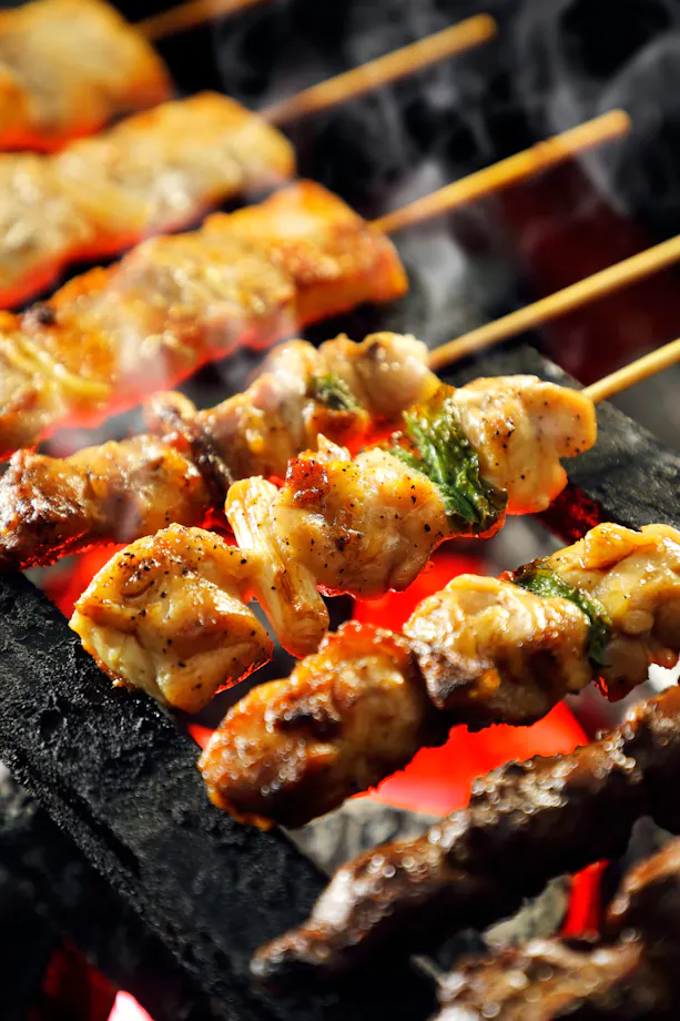 Skewers of grilled meat and vegetables cook over a charcoal grill, with visible smoke and red-hot embers below, creating a delicious, smoky atmosphere.