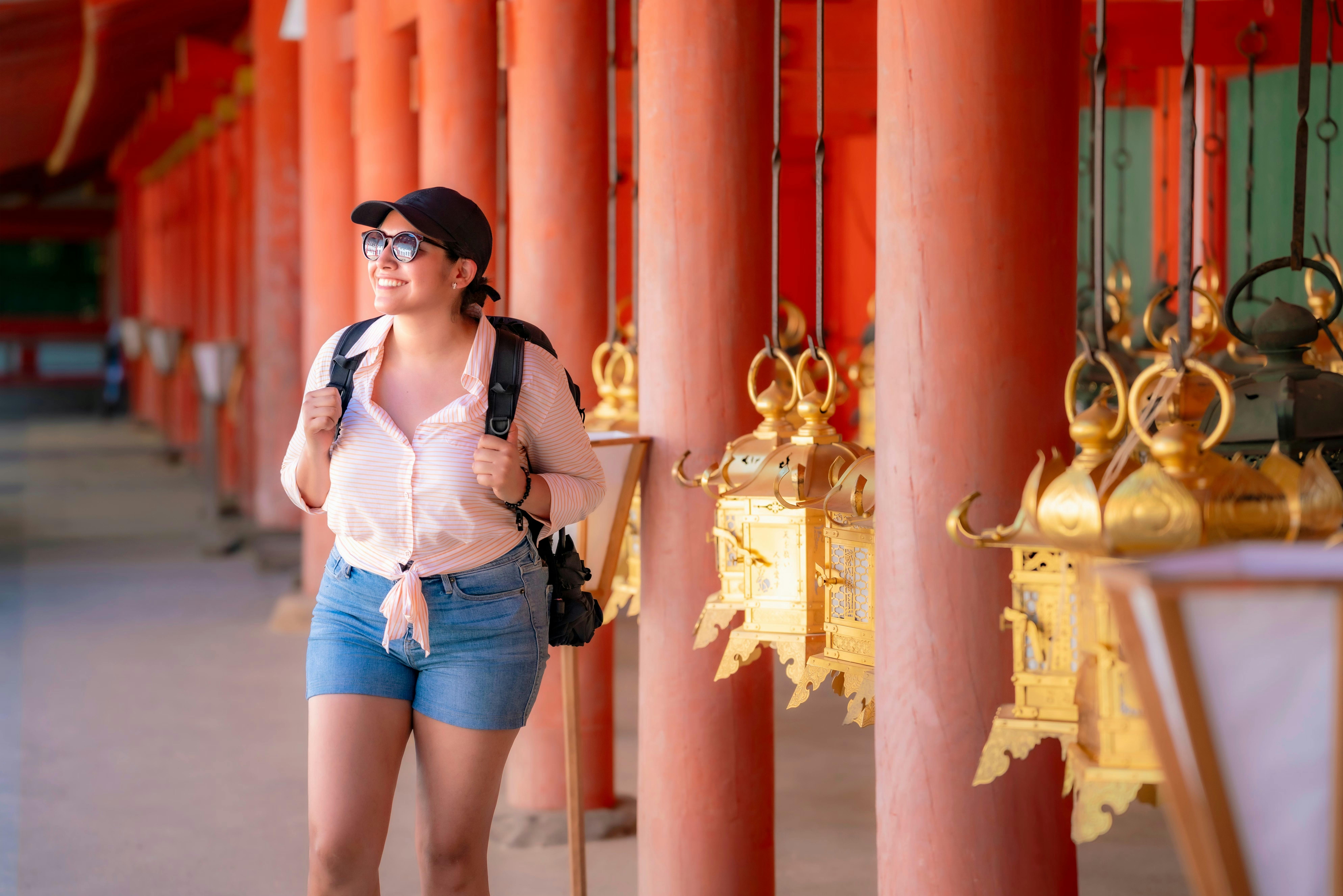 A smiling woman wearing sunglasses, a cap, and a backpack walks past ornate golden lanterns hanging from red pillars inside a traditional building, likely a temple. Sunlight illuminates the scene.