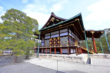A traditional Japanese wooden building with a sloped roof and ornate gold details, surrounded by gravel paths and a large green pine tree, under a bright blue sky.