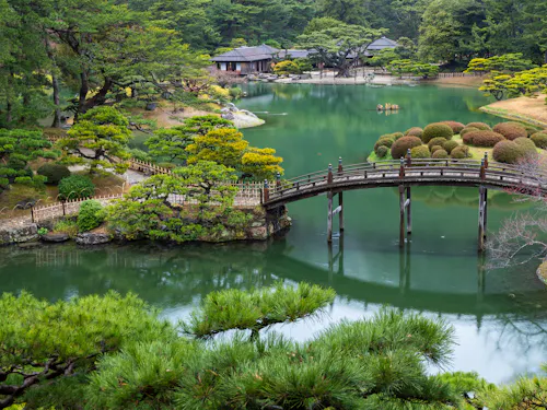 Takamatsu A traditional wooden bridge arches over a tranquil pond surrounded by manicured trees and shrubs in a serene Japanese garden, with wooden buildings visible in the background.