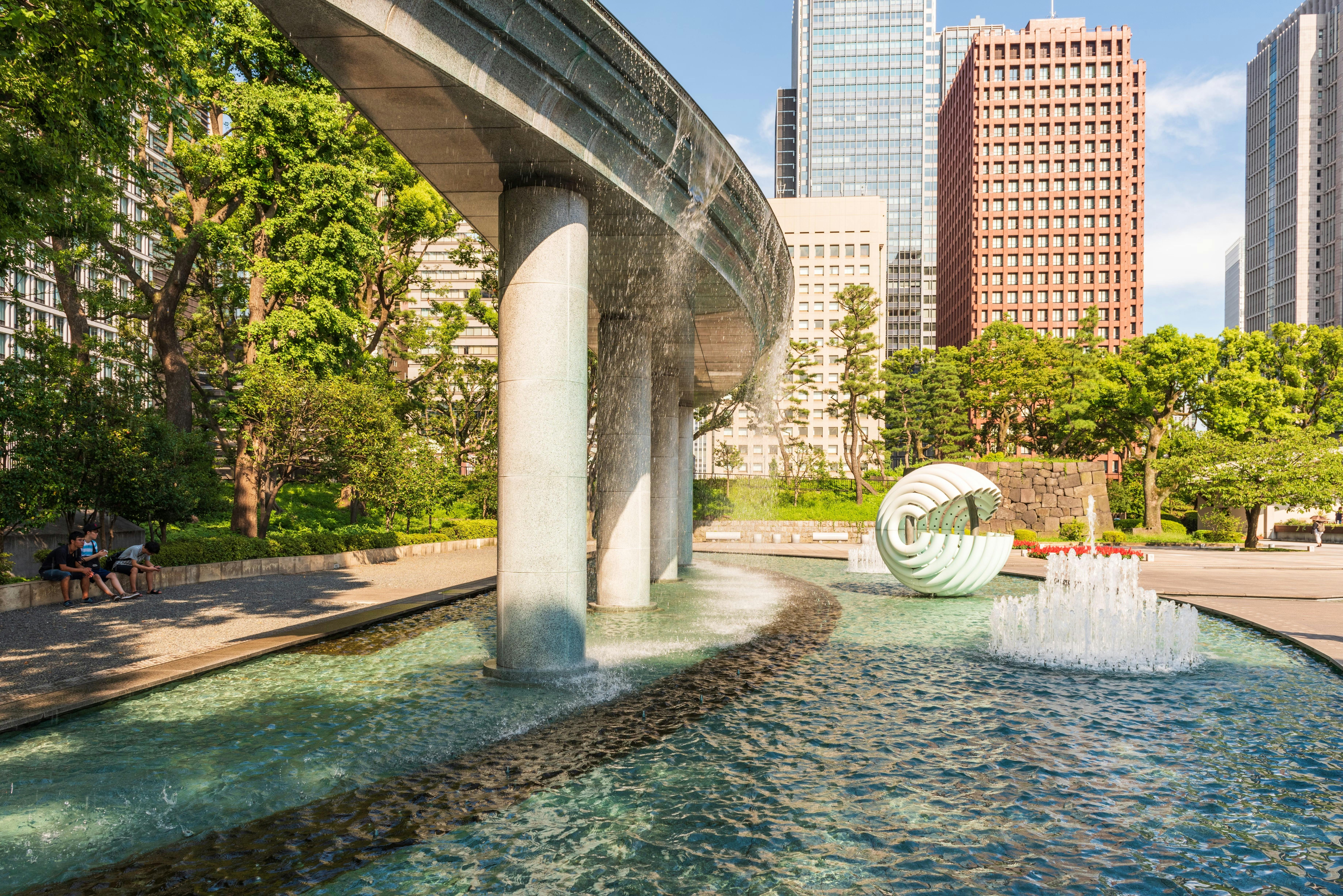 A modern city park features a curved elevated monorail, a fountain, a spiral sculpture, and surrounding trees, with tall office buildings in the background under a clear blue sky.