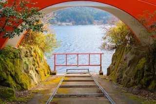 A view through an orange archway framing a path with old tracks, ending at a barrier by a calm lakeshore, surrounded by mossy rocks and greenery. A forest and lakeside buildings are visible in the background.