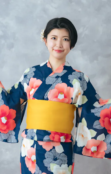 Wearing Kimono A woman wearing a blue floral kimono with a yellow obi poses with her arms spread, smiling in front of a light gray textured background.