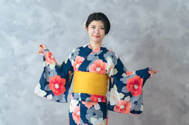 A woman wearing a blue floral kimono with a yellow obi poses with her arms spread, smiling in front of a light gray textured background.