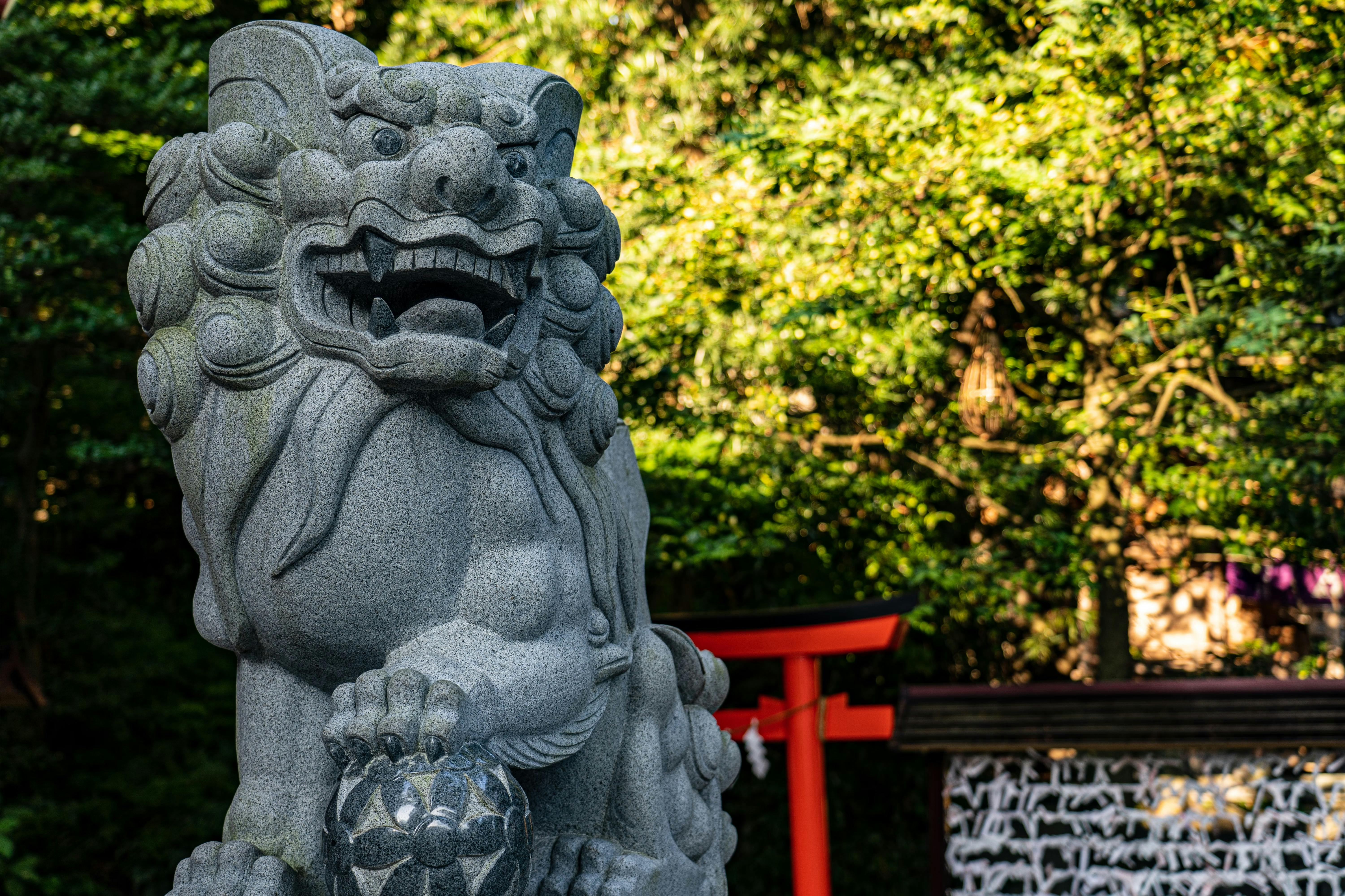 A stone statue of a lion guardian, known as komainu, stands outdoors near a red torii gate, with a background of green trees and sunlight filtering through the leaves.