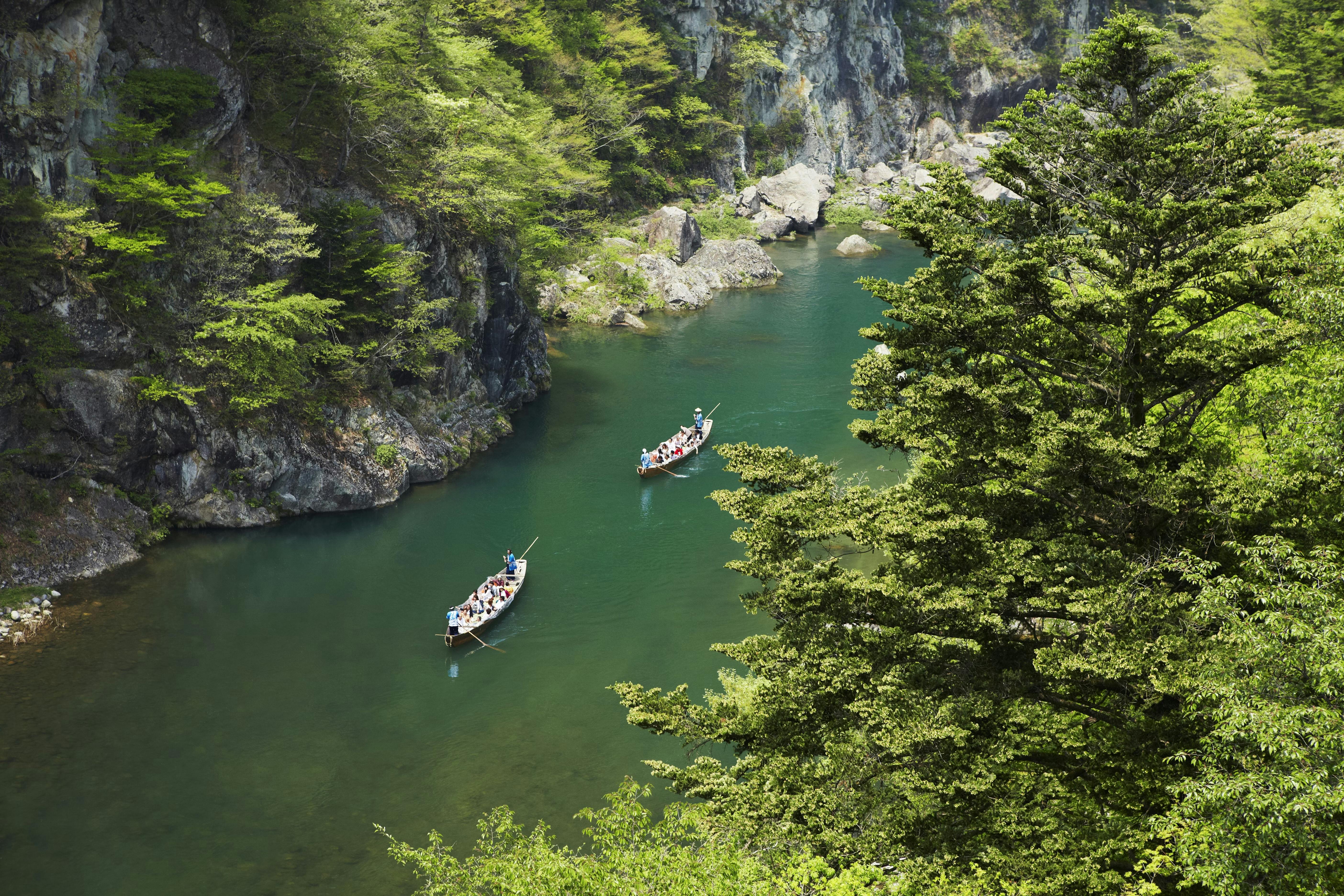 Two boats with people row along a river surrounded by steep cliffs and lush green trees in a scenic, forested canyon.
