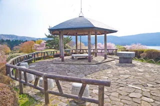 A wooden gazebo with a stone bench stands on a stone-paved platform surrounded by a wooden fence, with blooming trees and mountains in the background.