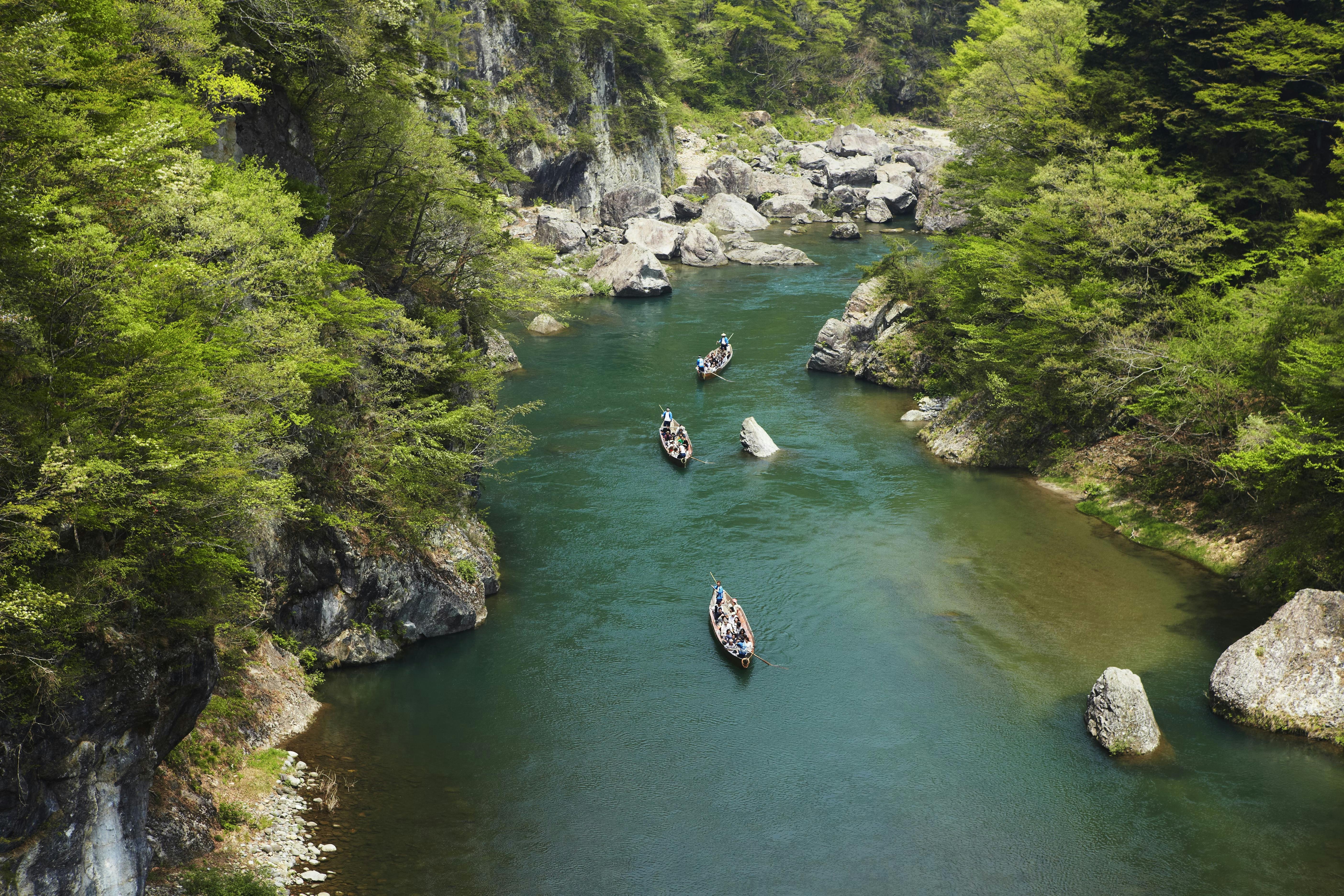 A river winds through a forested canyon, with several wooden boats carrying people gently floating downstream between rocky banks and lush green trees.