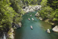 A river winds through a forested canyon, with several wooden boats carrying people gently floating downstream between rocky banks and lush green trees.