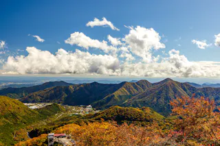 A scenic view of rolling mountains covered in autumn foliage under a bright blue sky with scattered clouds, with some buildings visible in the lower left corner.