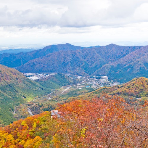 Akechidaira Observation Area A scenic mountain landscape in autumn, with rolling hills covered in vibrant red, orange, and yellow foliage under a cloudy sky. A small village is visible in the valley below.