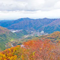 Akechidaira Observation Area A scenic mountain landscape in autumn, with rolling hills covered in vibrant red, orange, and yellow foliage under a cloudy sky. A small village is visible in the valley below.