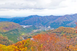 A scenic mountain landscape in autumn, with rolling hills covered in vibrant red, orange, and yellow foliage under a cloudy sky. A small village is visible in the valley below.
