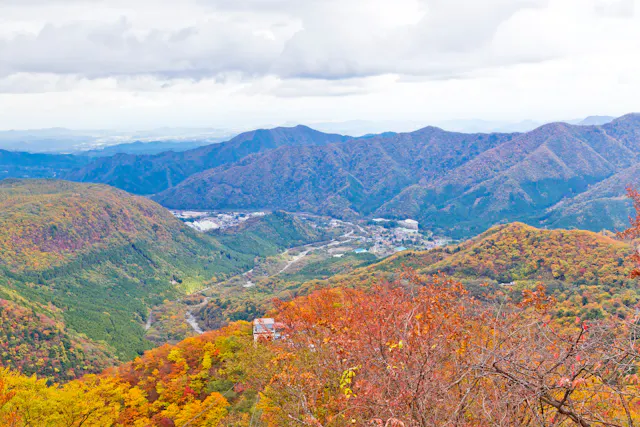 A scenic mountain landscape in autumn, with rolling hills covered in vibrant red, orange, and yellow foliage under a cloudy sky. A small village is visible in the valley below.