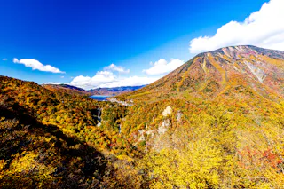 A vibrant autumn landscape with colorful foliage covering hills and mountains under a bright blue sky; a waterfall flows into a river in the valley, and a distant lake is visible.