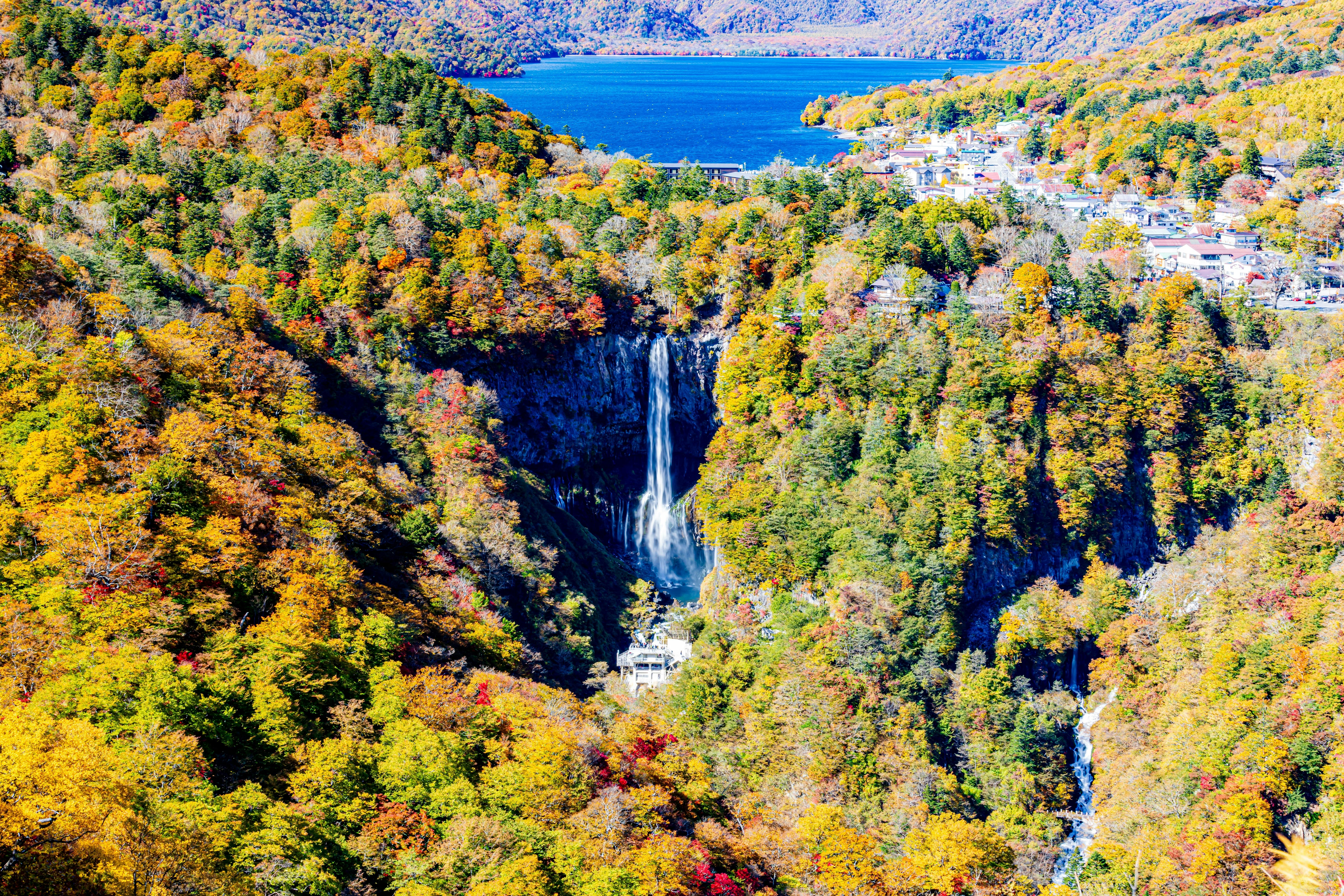 A tall waterfall cascades through a forest of vibrant autumn foliage, with a blue lake and a small town visible in the background under a clear sky.