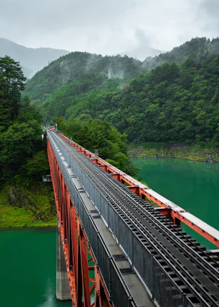 A red railway bridge stretches over a calm, green river, surrounded by lush green forested hills and misty mountains under an overcast sky.