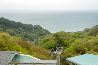 A scenic view of lush green hills with a cable car tower in the foreground, leading down to the ocean in the background under a cloudy sky.