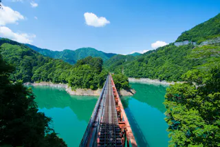 A red railway bridge stretches across a turquoise river surrounded by lush green mountains under a bright blue sky with a few clouds.