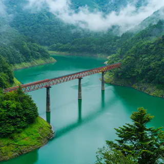 A red railway bridge crosses over a turquoise river surrounded by lush green forest and misty mountains. The scene is tranquil and vibrant, with clouds hovering above the landscape.