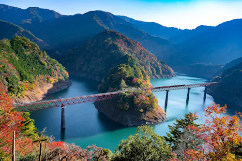 A red bridge spans a turquoise river surrounded by forested mountains with autumn foliage, under a clear blue sky. The scene is peaceful and vibrant with fall colors.
