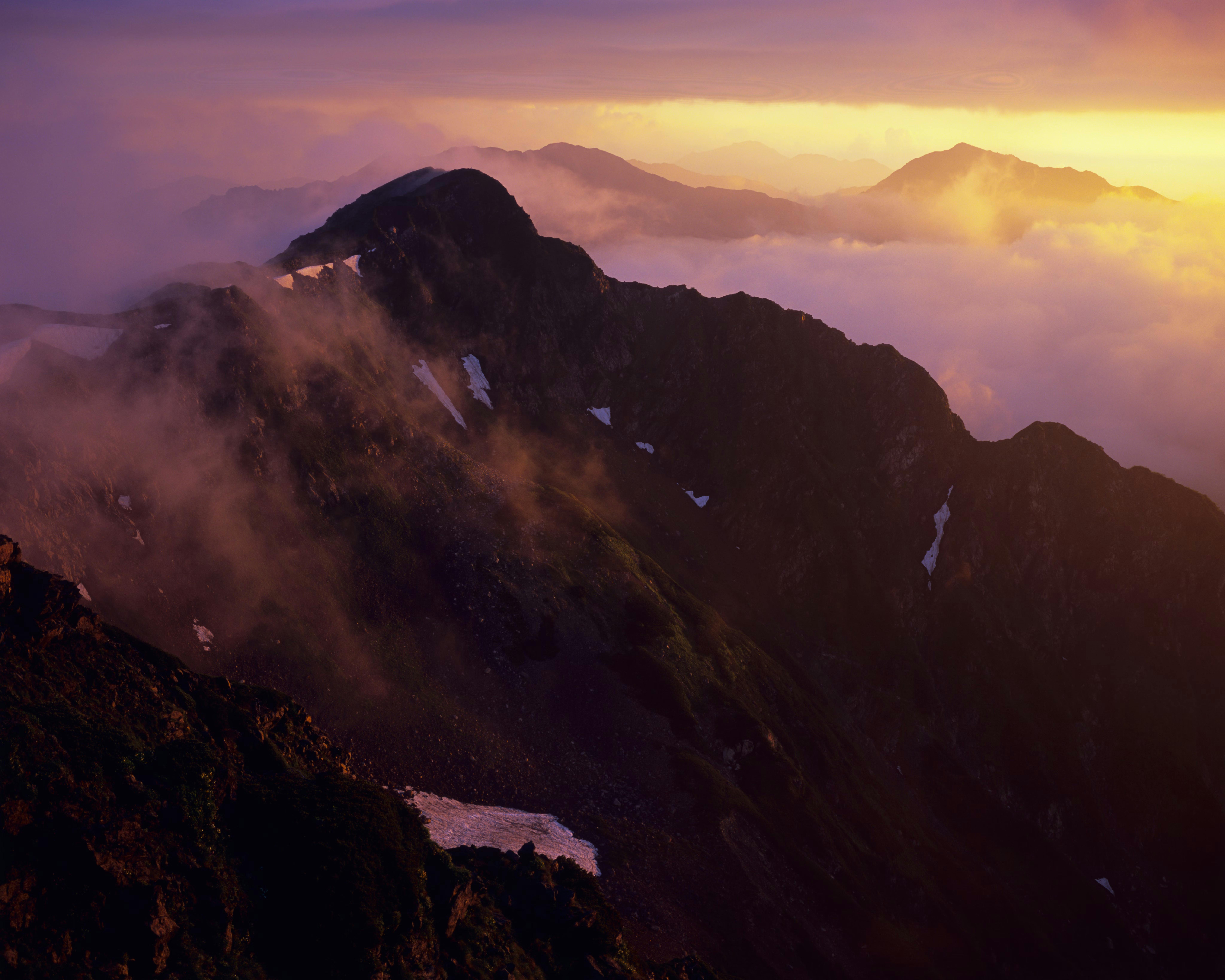 A mountain ridge is covered in patches of snow and mist, with soft clouds and layers of distant peaks glowing in the warm, golden light of sunrise or sunset.