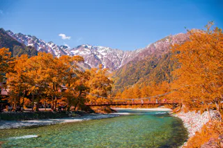 A wooden bridge crosses a clear river, surrounded by trees with vibrant orange autumn leaves, set against a backdrop of tall, snow-capped mountains under a bright blue sky.