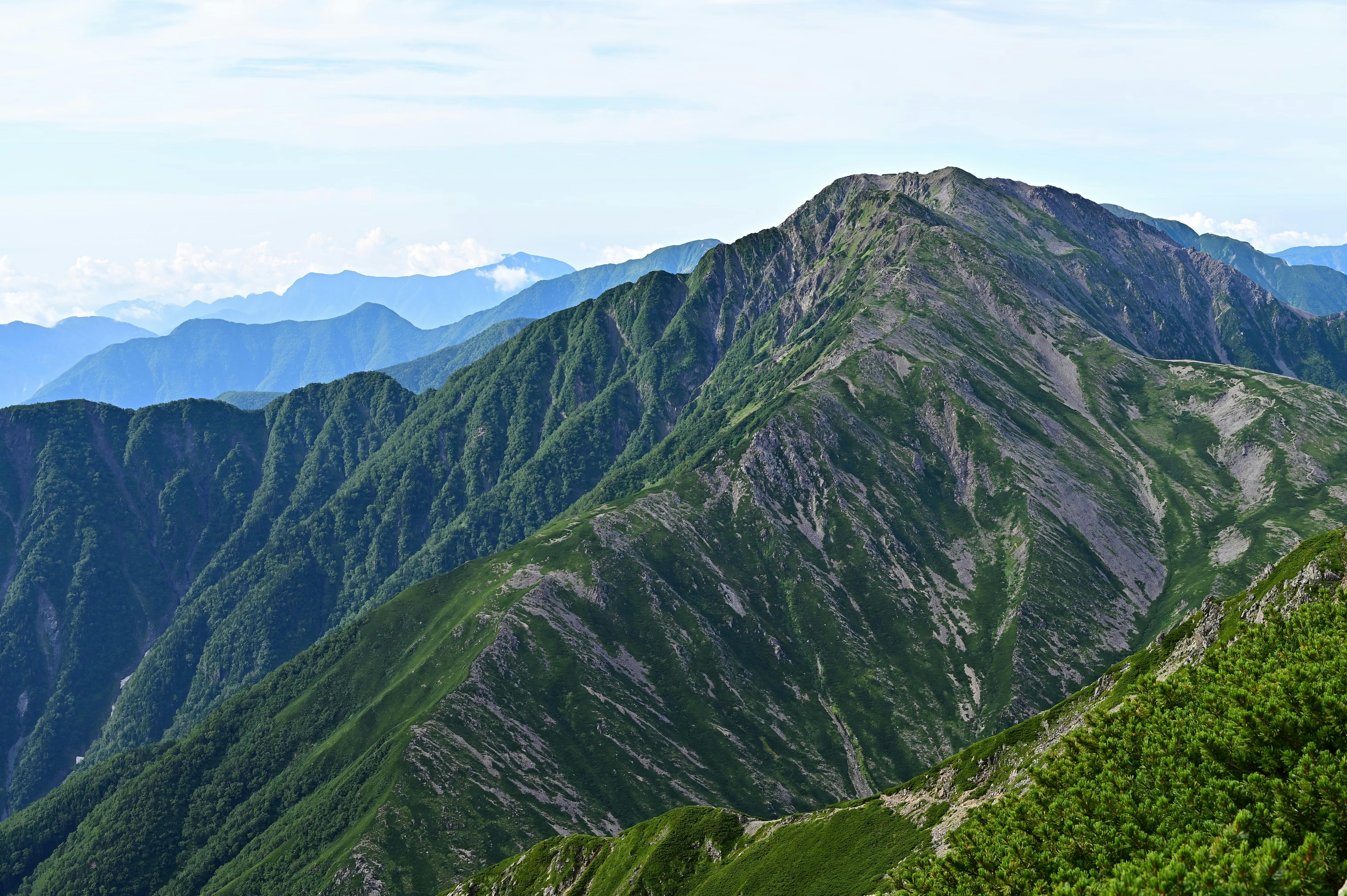 A rugged mountain ridge covered in green vegetation stretches into the distance under a partly cloudy sky, with overlapping mountain ranges visible on the horizon.