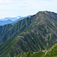 Mount Akaishi A rugged mountain ridge covered in green vegetation stretches into the distance under a partly cloudy sky, with overlapping mountain ranges visible on the horizon.