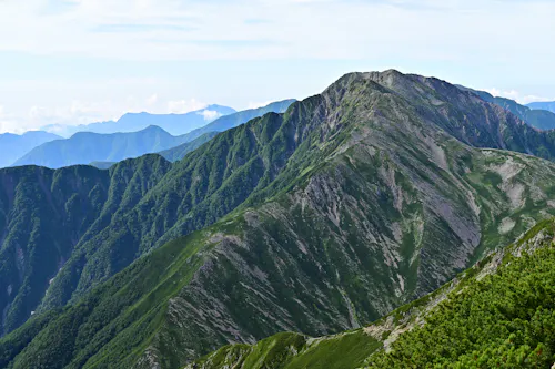 A rugged mountain ridge covered in green vegetation stretches into the distance under a partly cloudy sky, with overlapping mountain ranges visible on the horizon.