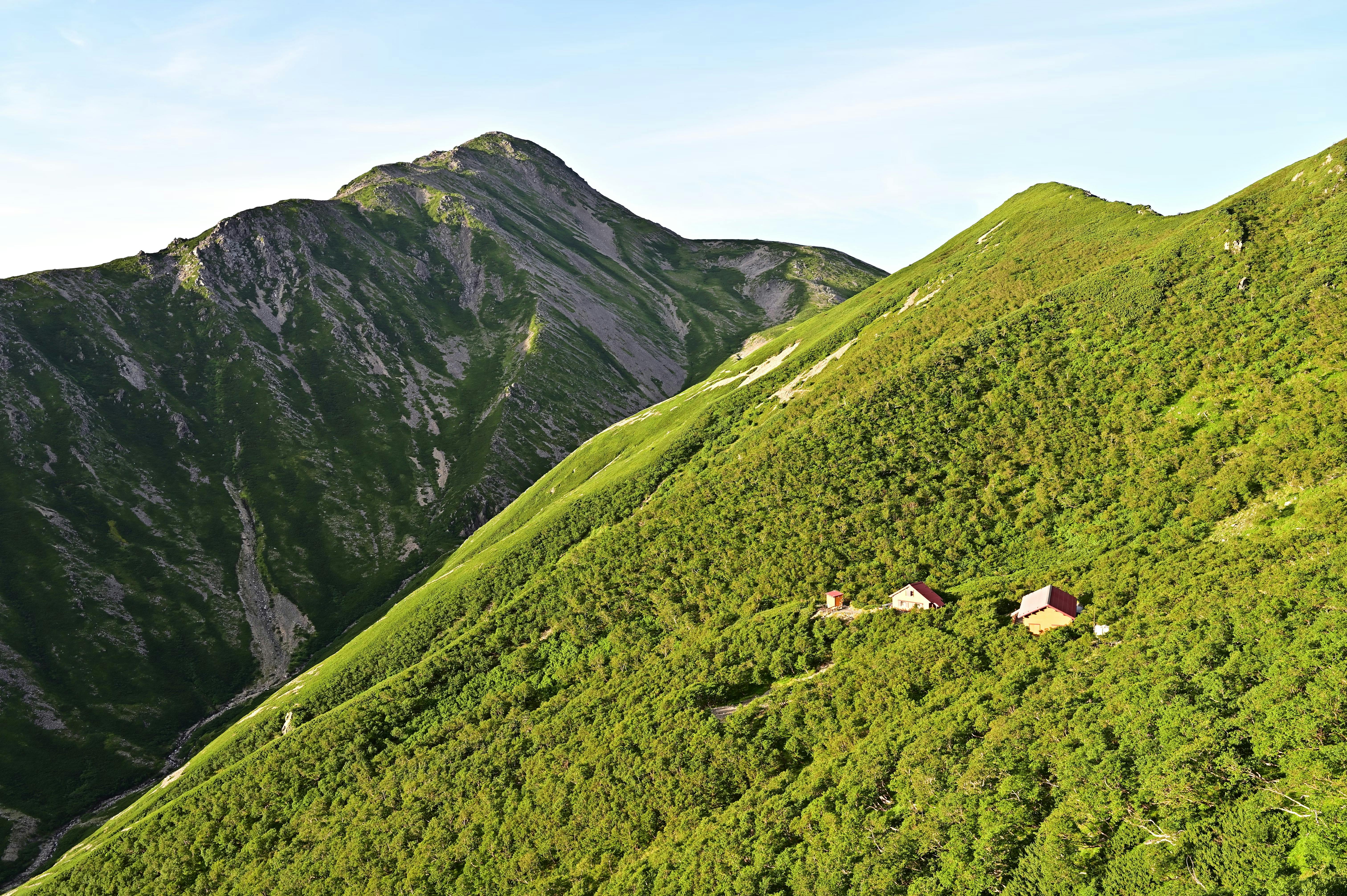 Small houses sit nestled among lush green hills, with a steep, rugged mountain rising in the background under a clear sky.