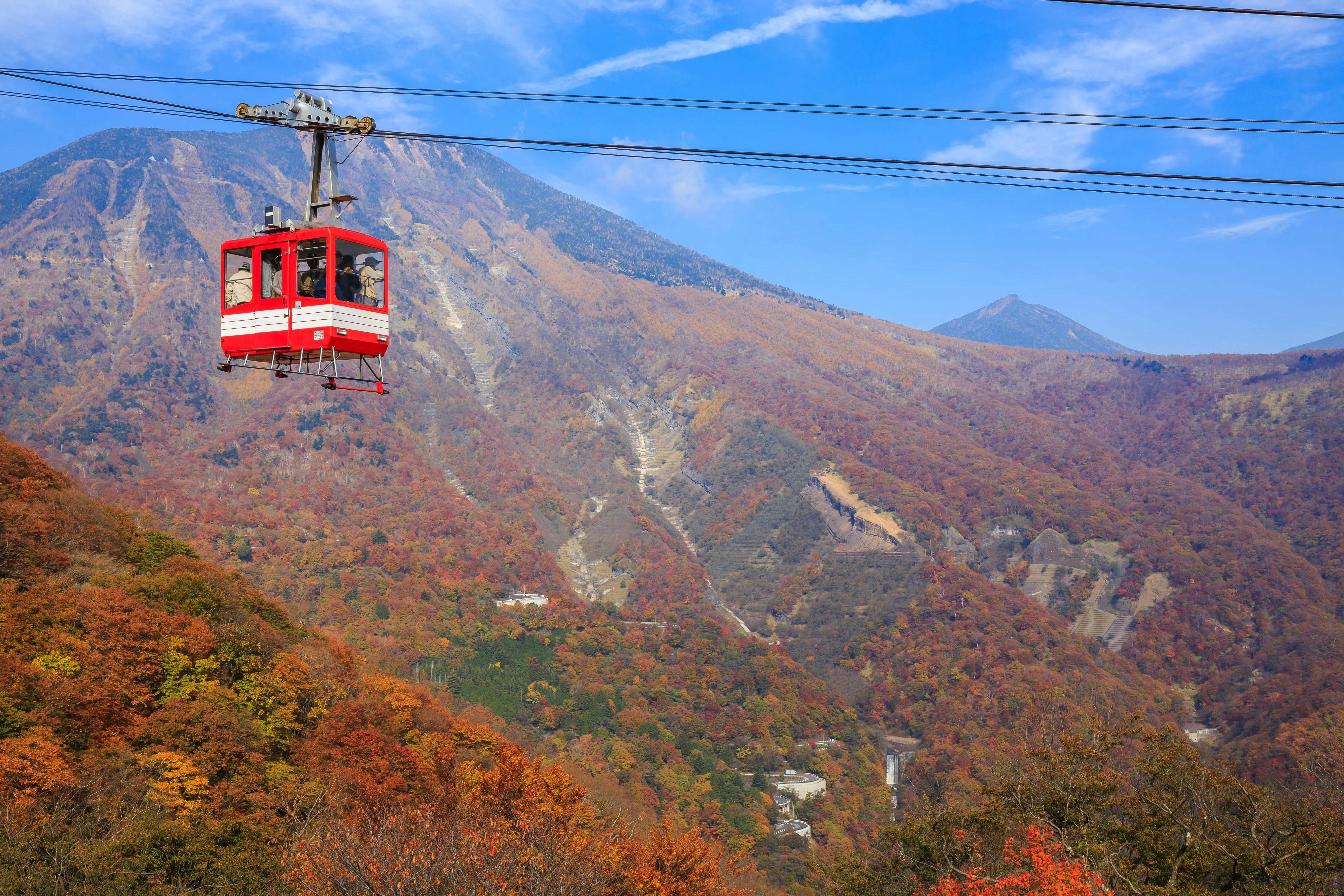 A red and white cable car travels over a forested mountain landscape with autumn foliage under a bright blue sky with wispy clouds.