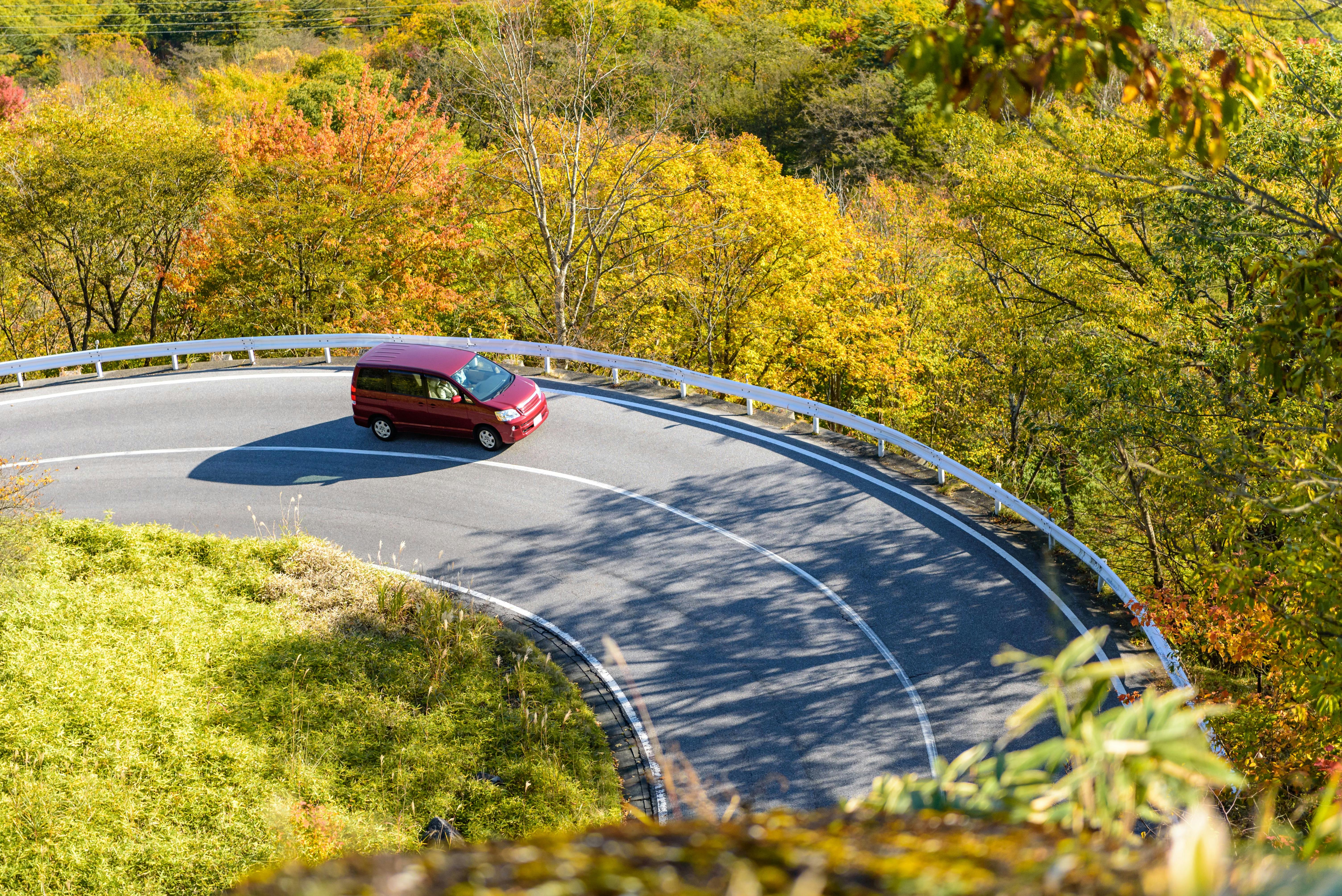 A red car drives around a sharp curve on a paved mountain road, surrounded by vibrant autumn trees with yellow, orange, and green foliage. The scene is sunny and bright.