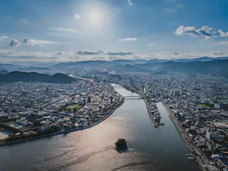 Aerial view of a city divided by a wide river, with a small green island in the water, surrounded by buildings, mountains in the distance, and the sun shining in a blue sky with scattered clouds.
