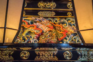 Colorful carved dragon with red and gold details surrounded by swirling blue waves, displayed on a glossy black and gold ornamental wooden structure, partially framed by two lanterns.
