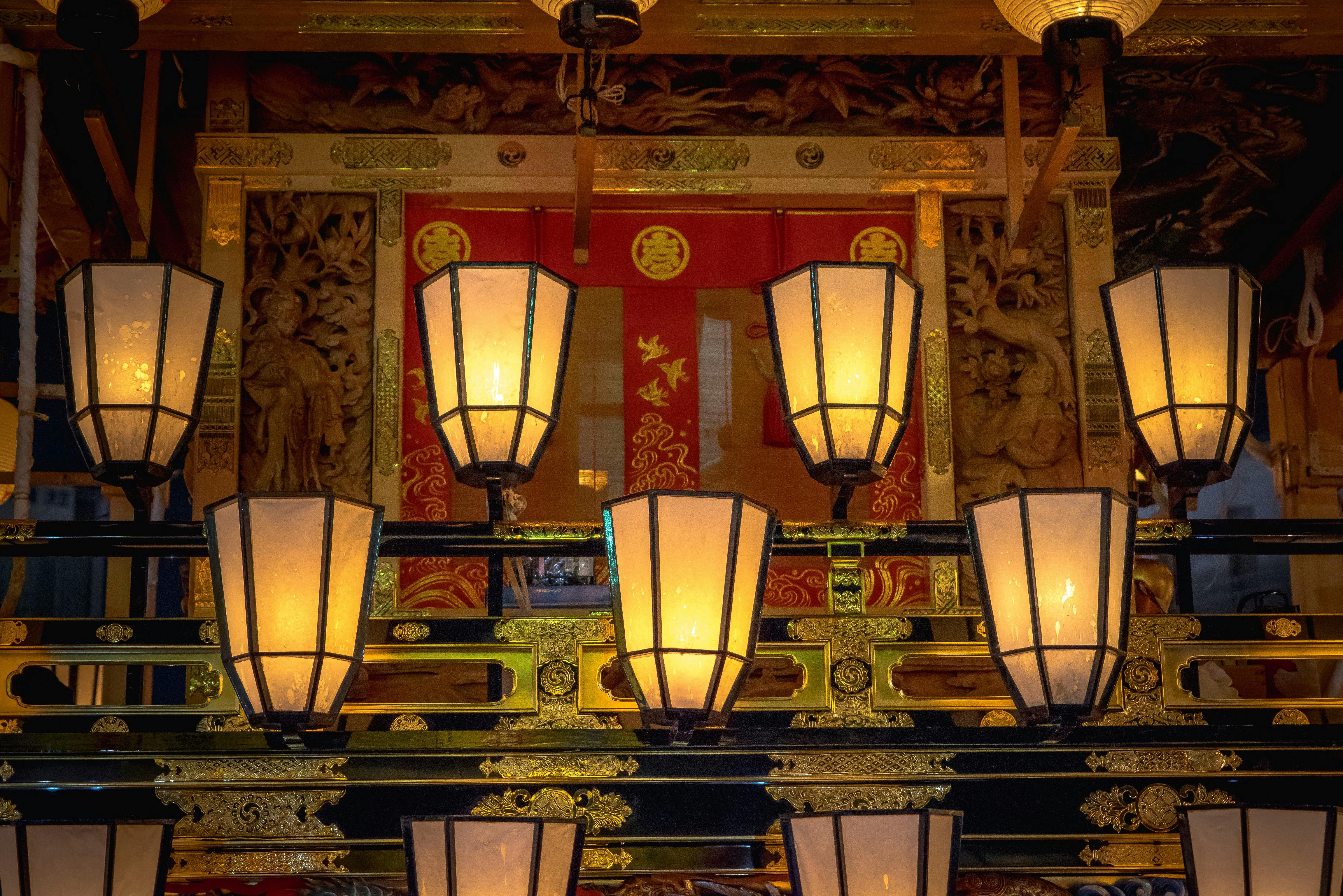 Rows of glowing lanterns illuminate an ornate, gold-decorated structure with intricate carvings and a red curtain in the background.
