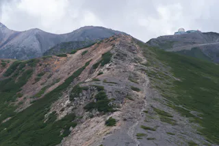 A narrow, rocky mountain ridge with patches of green vegetation stretches toward distant peaks under a cloudy sky. A small building or observatory is visible on a nearby slope.