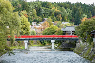 A bright red bridge spans a gently flowing river, surrounded by lush green trees and houses nestled on a hillside in the background.