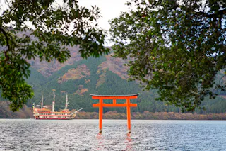 A bright red torii gate stands in a lake, framed by green tree branches. In the background, a pirate-style sightseeing boat sails with forested hills and mountains rising beyond the water.