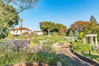 A landscaped garden with colorful flowers, trimmed bushes, a curved pathway, and a white house with a red roof in the background, set against a bright blue sky and lush trees.