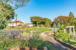 A landscaped garden with colorful flowers, trimmed bushes, a curved pathway, and a white house with a red roof in the background, set against a bright blue sky and lush trees.