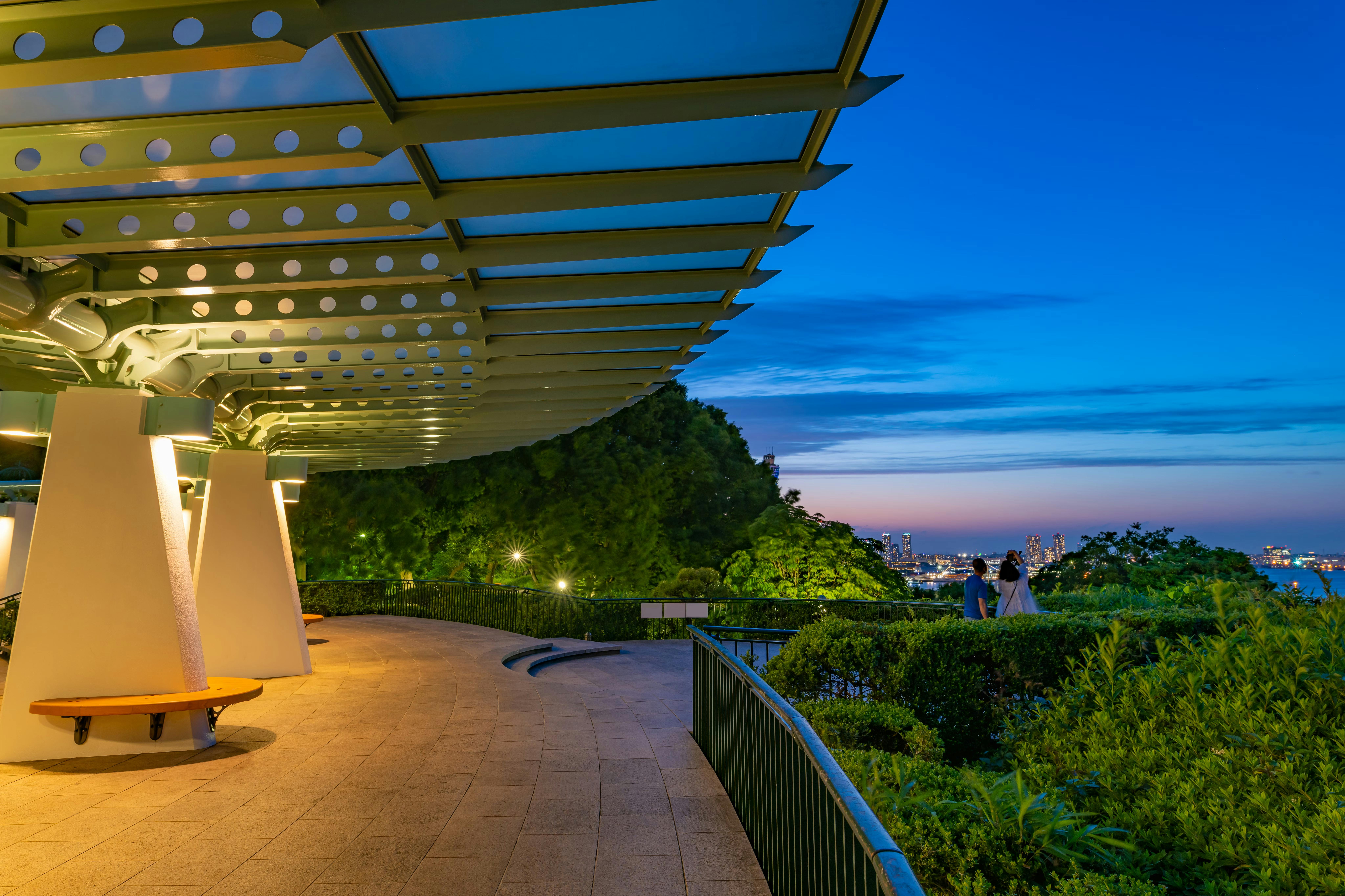 Covered walkway with modern metal roof, lit by yellow lights, curves along lush greenery. Two people stand by a railing, overlooking a cityscape at twilight under a deep blue sky.