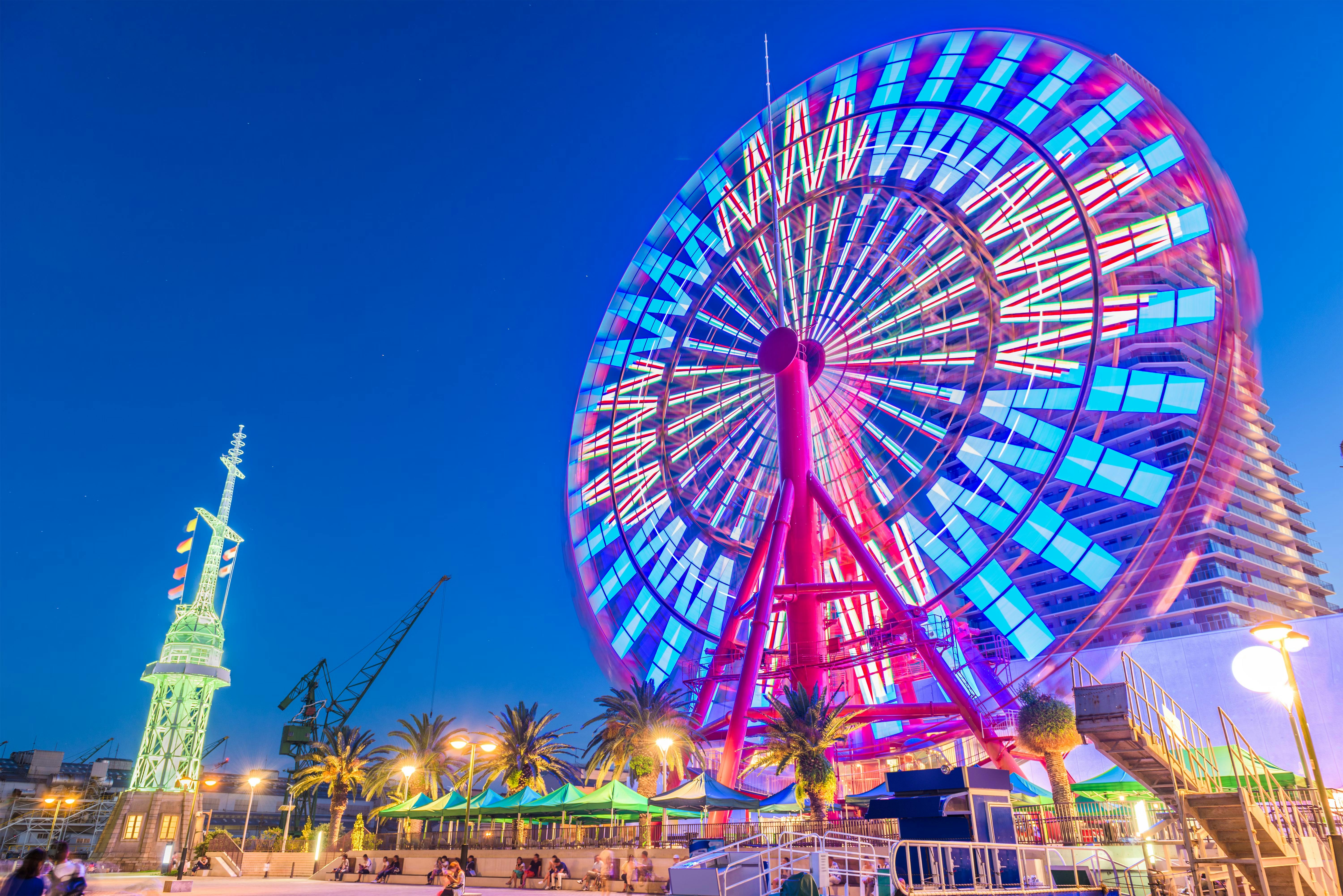 A large, colorful Ferris wheel with vibrant LED lights glows at night near palm trees and a lit tower, set against a clear blue evening sky in an urban waterfront area.