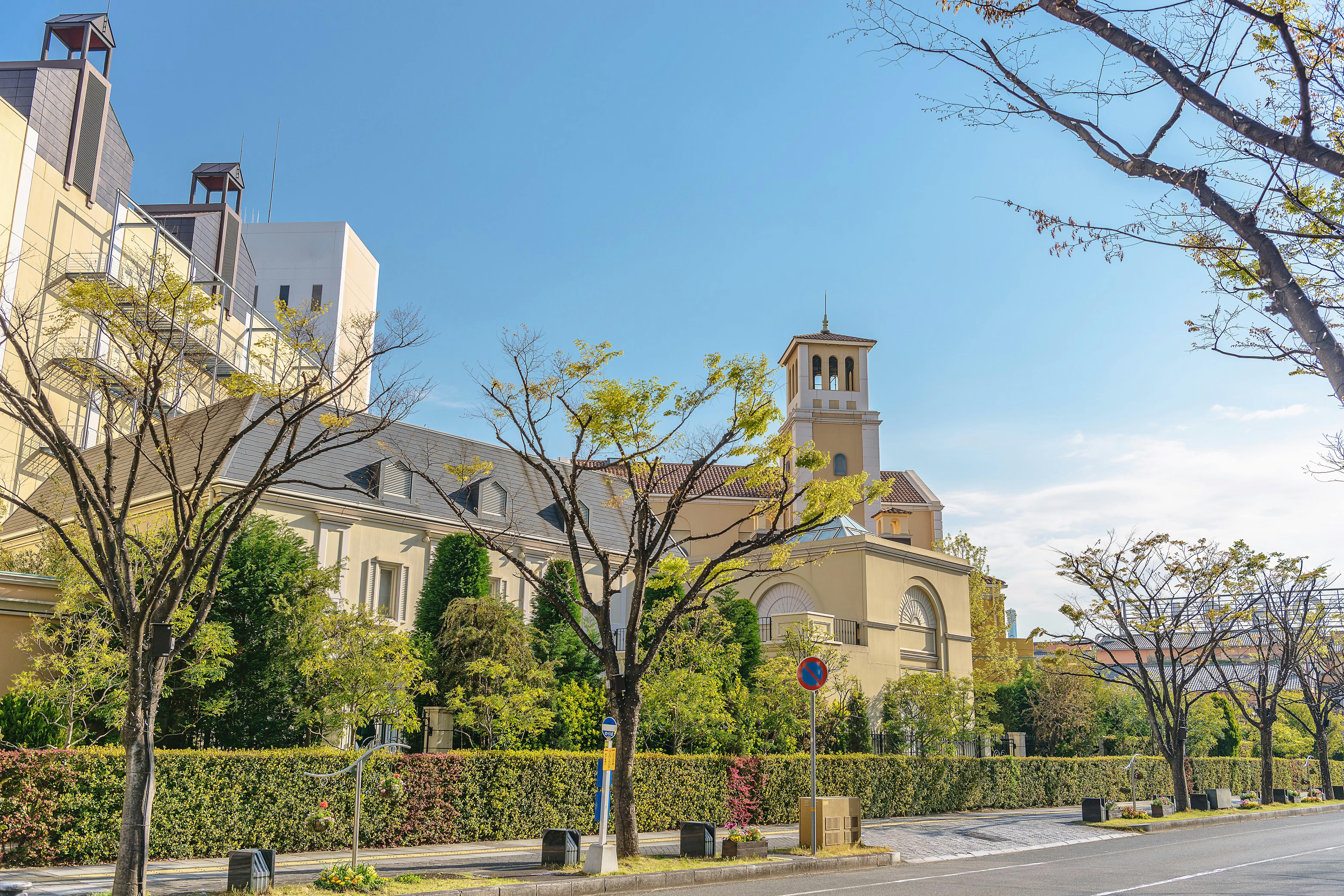 A tree-lined street with a beige building featuring a tower and arched windows, surrounded by green hedges and shrubs under a clear blue sky.