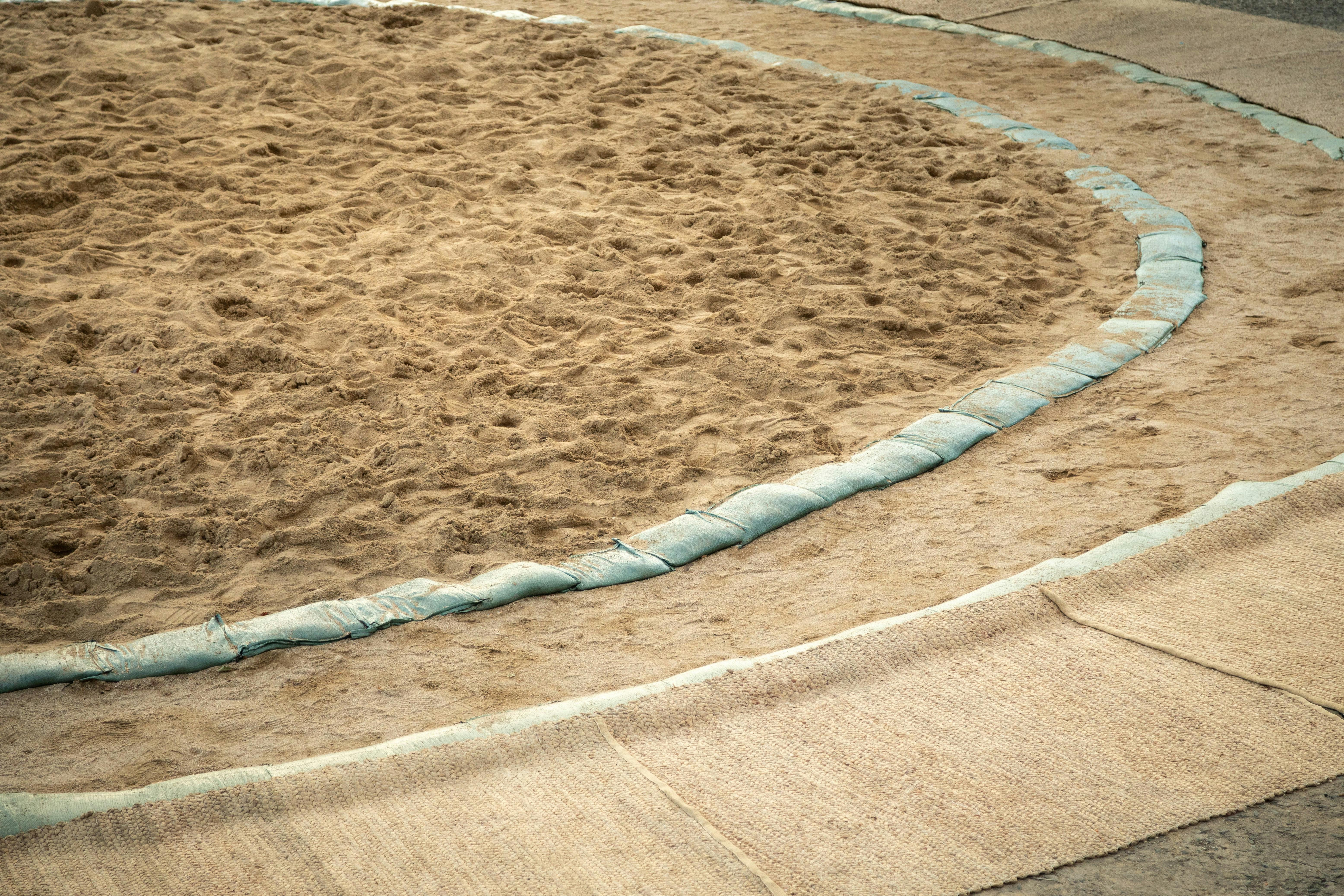 A close-up view of a sumo wrestling ring, featuring a circular area of packed sand bordered by greenish sandbags and straw mats on the outside.