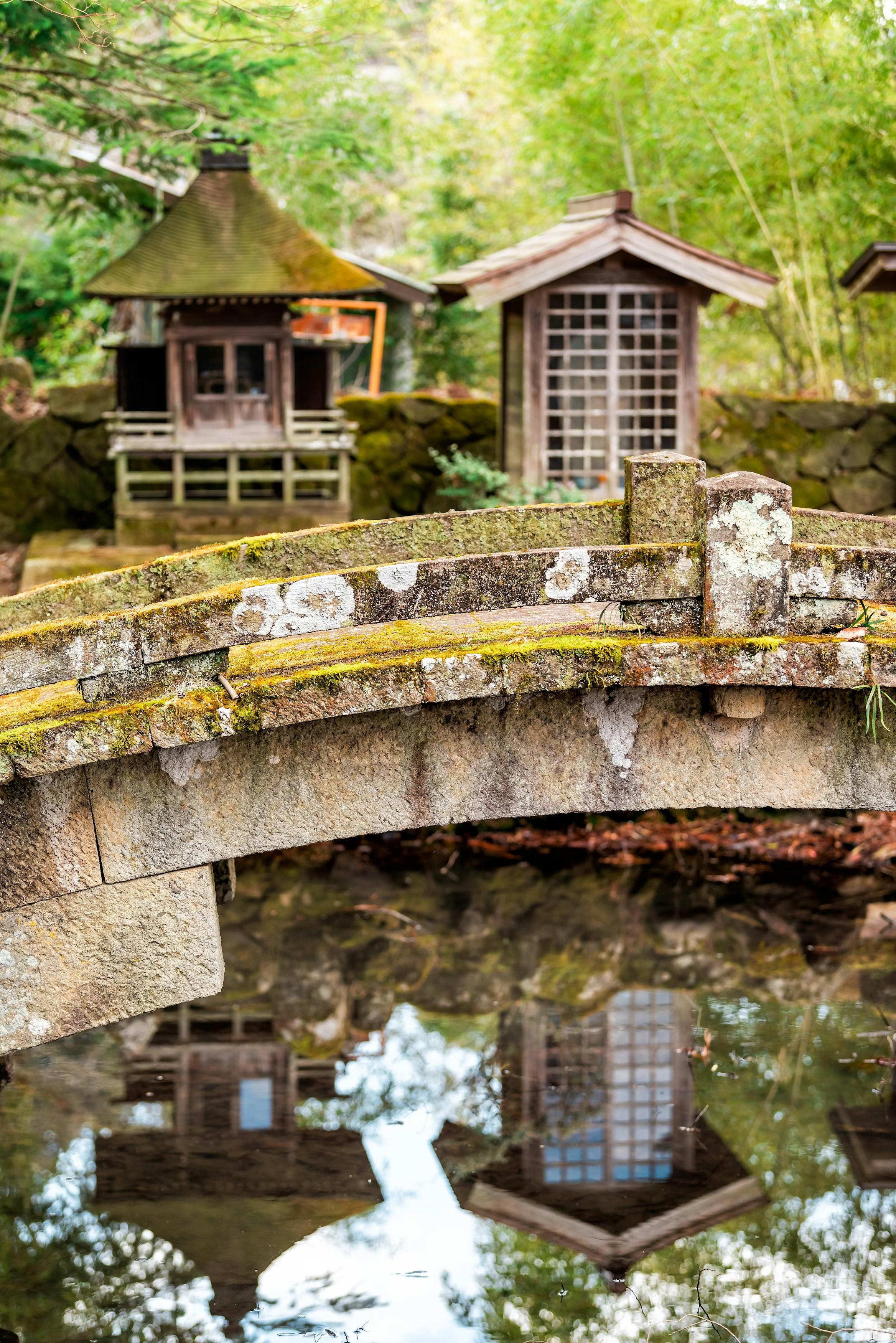 A stone bridge covered in moss crosses over a pond, reflecting two small wooden Japanese-style buildings surrounded by lush greenery in a tranquil garden setting.