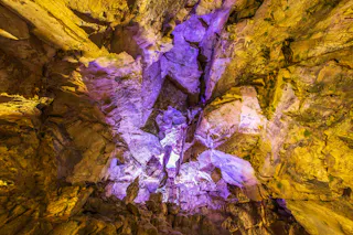 A rocky cave ceiling illuminated with vibrant yellow and purple lights, highlighting rough stone textures and jagged formations.