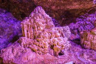 A large, textured rock formation inside a cave is illuminated by purple lighting, highlighting its jagged and layered surfaces. The cave walls surround the formation, creating a vibrant underground scene.