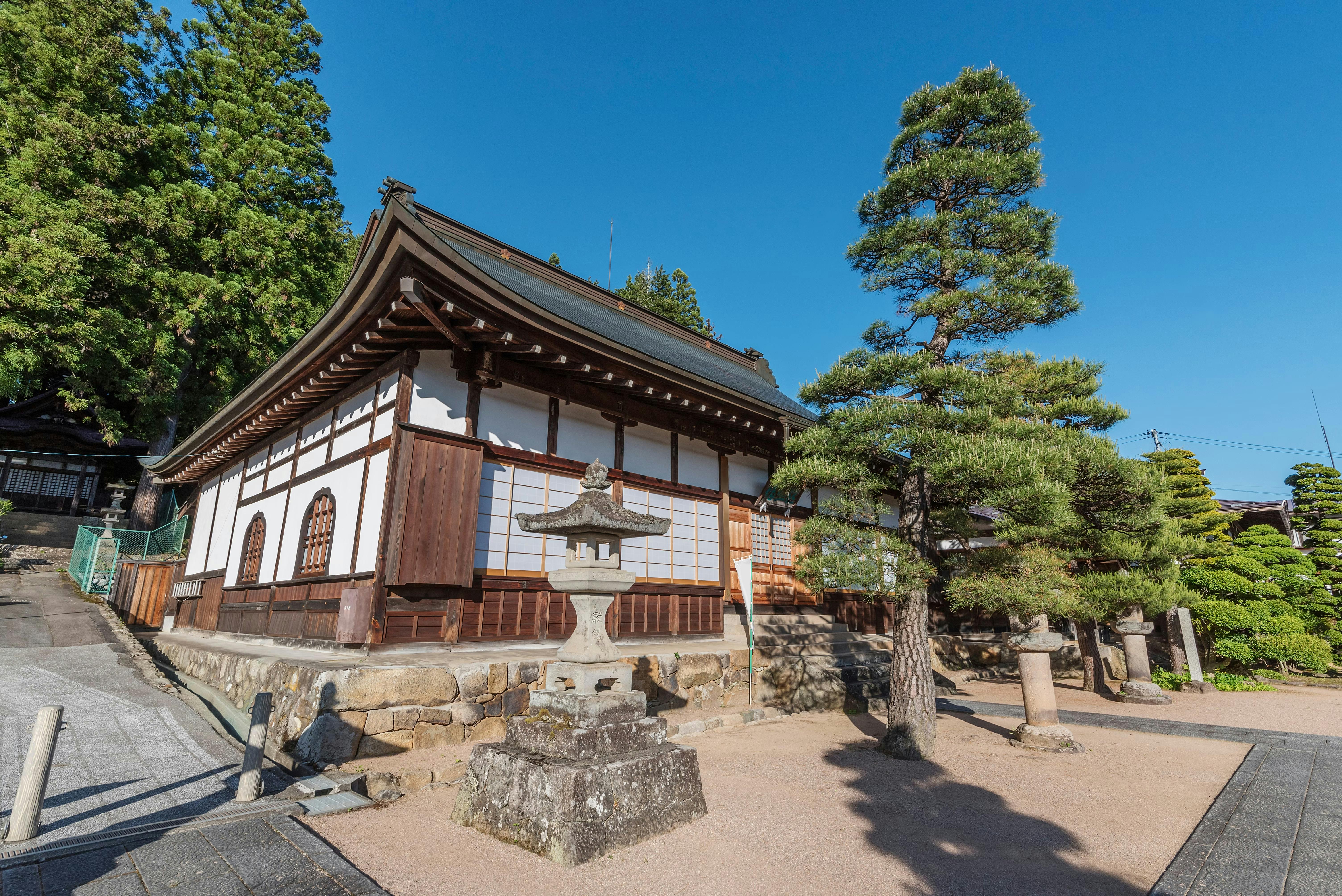 Traditional Japanese temple building with wooden beams and white walls, surrounded by neatly trimmed trees and a stone lantern, under a clear blue sky.